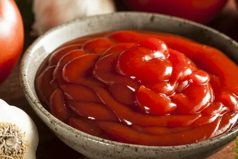 Bowl of fresh ketchup next to a fresh tomato and garlic bulb