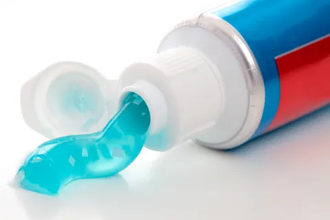 Open tube of toothpaste laying on a white table with teal translucent gel coming out 